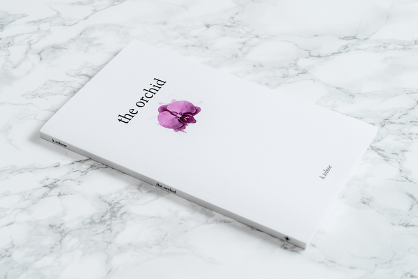 An angled shot of the front of "the orchid" poetry book, featuring the title, the author name k.tolnoe and a watercolor image of an orchid on a marble background.